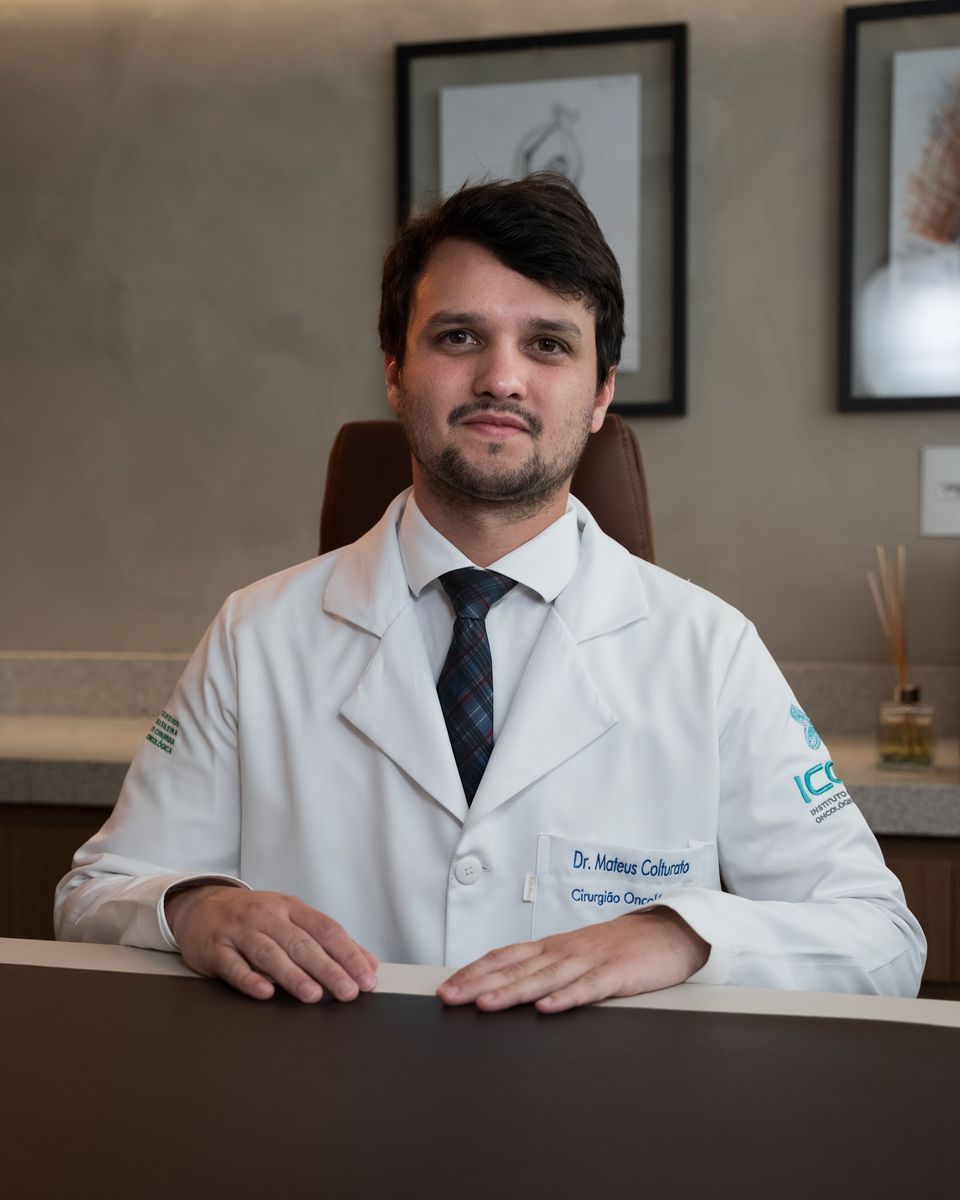 Dr. Mateus Colturato de jaleco, sentado à mesa do consultório